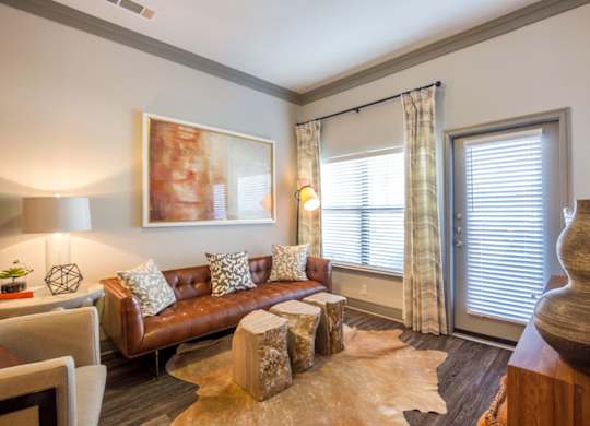 A living room here at Sorrel Fairview with a warm brown leather sofa, light patterned pillows, beige accent chair, tan hide rug, wood stump tables, and soft neutral lighting by the window and balcony door.