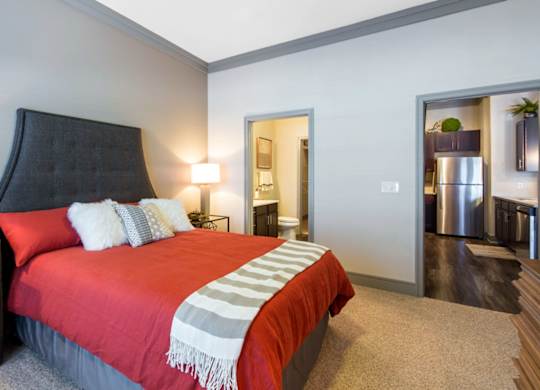 A bedroom here at Sorrel Fairview with a dark gray tufted headboard, red bedspread, white accent pillows, striped throw blanket, soft carpet flooring, and warm bedside lamp lighting.