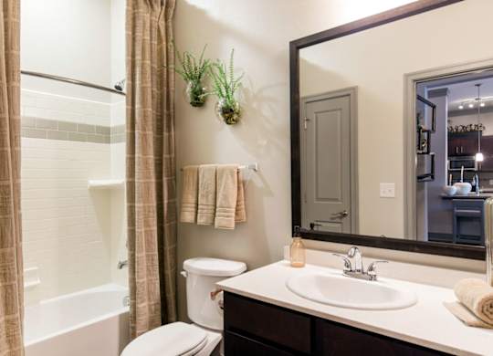 A bathroom here at Sorrel Fairview with a white tub-shower combo and beige curtain, dark wood vanity, wide framed mirror, neutral countertop, tan towels, and small green wall plants for a warm, clean look.