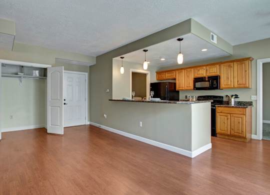 A bright open living and kitchen space here at Bridges with warm wood flooring, natural wood cabinets, a breakfast bar with pendant lights, black appliances, and an open entry closet near the front door.