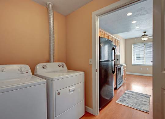 A laundry area here at Bridges with a white washer and dryer, peach-toned walls, and a vent duct, opening into a bright kitchen with wood cabinets, black refrigerator, and warm wood-look flooring.