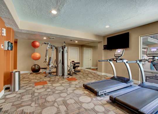 A fitness room here at Bridges with treadmills, a cable weight machine, wall mirrors, mounted TV, patterned carpet, and orange accent walls with exercise balls and strength equipment.