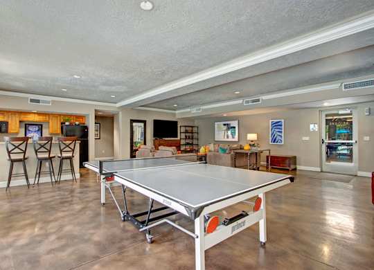 A game and lounge room here at Bridges with a ping pong table, kitchen bar with wood cabinets and stools, seating area with sofas and TV, and polished concrete floors in a bright open layout.