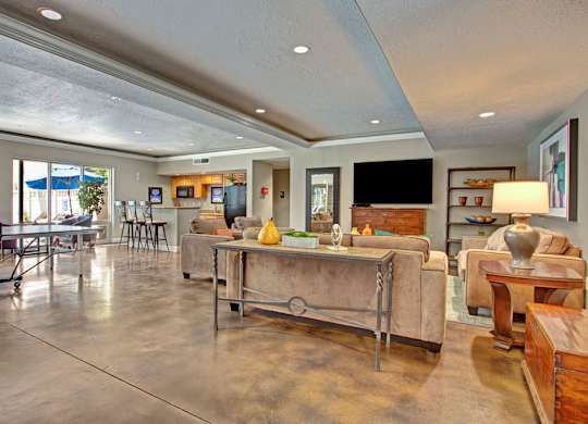 A game and lounge room here at Bridges with tan sofas, wood accent tables, ping pong table, bar-height kitchen counter with stools, TV wall, and polished concrete floors in a bright, open layout.