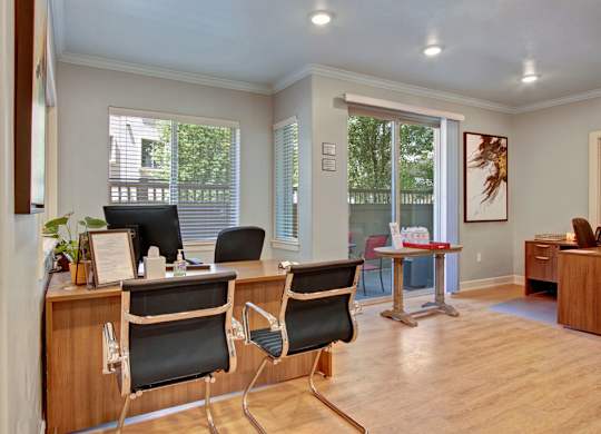 A leasing office here at Bridges with warm wood desks, black guest chairs, light wood floors, and bright windows with patio view, creating a clean, welcoming workspace.