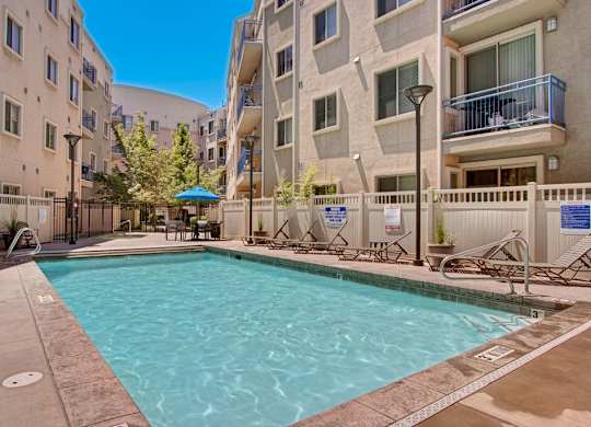 A pool courtyard here at Bridges with a rectangular pool, beige apartment buildings, blue balcony railings, tan deck, and lounge chairs, creating a bright, inviting outdoor amenity space.