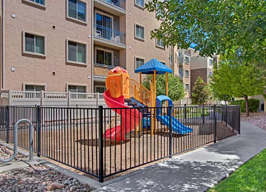 A playground courtyard here at Bridges with a colorful red and blue slide set, black safety fence, green trees, and surrounding apartment balconies, creating a bright, family-friendly outdoor space.