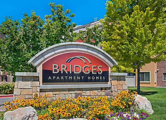 A community monument sign here at Bridges with a stone base and curved red-and-black Bridges Apartment Homes panel, surrounded by bright yellow flowers, green trees, and landscaped greenery under a clear blue sky.