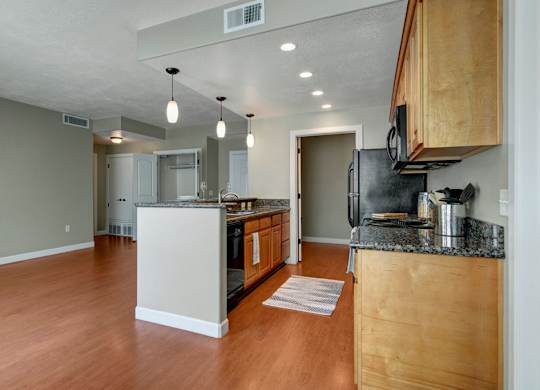 A bright open kitchen and living area here at Bridges with warm wood flooring, light wood cabinets, dark countertops, black appliances, a center island with pendant lights, and an open layout leading to nearby rooms.