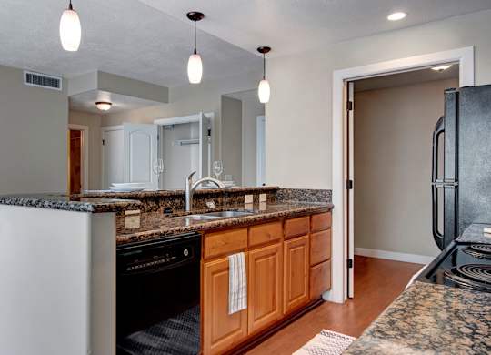 A kitchen here at Bridges with a granite breakfast bar and island sink, warm wood cabinets, black appliances, pendant lighting, and open sightlines to the living area and nearby rooms.