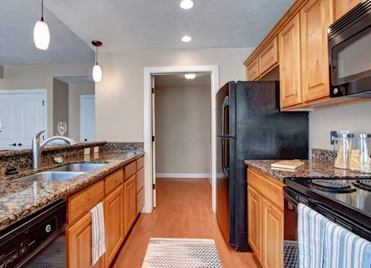 A kitchen here at Bridges with granite countertops, warm wood cabinets, black appliances, pendant lights, a double sink, and a long breakfast bar overlooking the open living space.