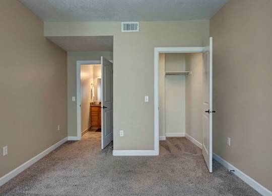 A bedroom here at Bridges with beige walls, gray carpet, an open white closet, and a doorway leading to a bright bathroom vanity area with warm wood cabinetry and mirror lighting.