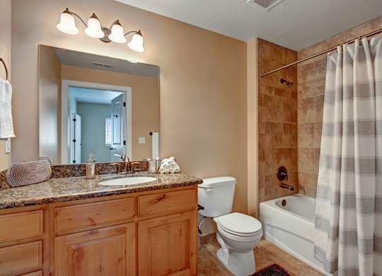 A bathroom here at Bridges with warm wood vanity cabinets, granite countertop, large mirror, white toilet, and a tub-shower combo with tan tile walls and a gray striped shower curtain under bright vanity lighting.