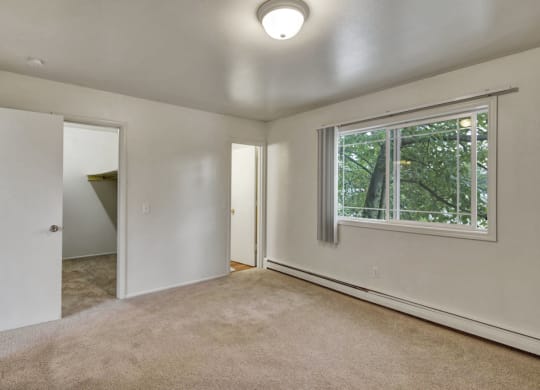 An empty bedroom here at the Village at Calais Apartments with beige carpet, white walls, and a window with vertical blinds showing green foliage outside. Open doors reveal a closet and another room.