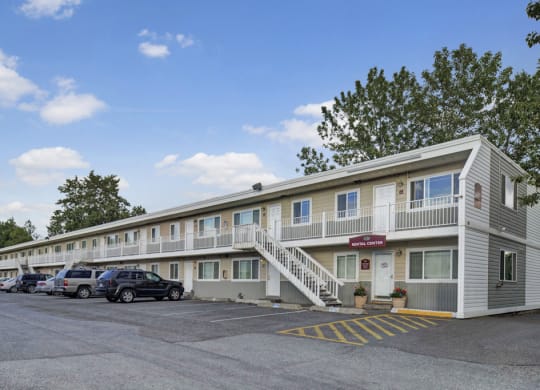 Two-story Village at Calais Apartment building with a staircase in the center, surrounded by parked cars. Clear blue sky, trees in the background; welcoming, calm atmosphere.