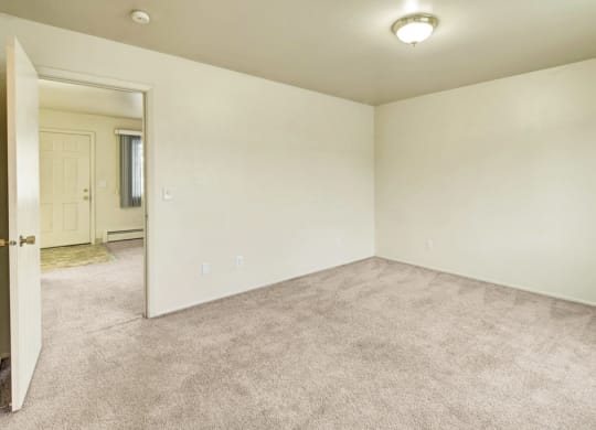 Village at CalaisEmpty beige room here at the Village at Calais Apartments with carpeted floor, plain walls, and an overhead light fixture. An open door reveals an adjacent space with a window and a closed front door. Minimalist and calm ambiance. Apartments - Livingroom