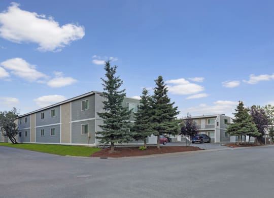 Two-story gray Village at Calais Apartment buildings with white trim, surrounded by lush trees and neatly manicured lawns under a bright blue sky with scattered clouds.