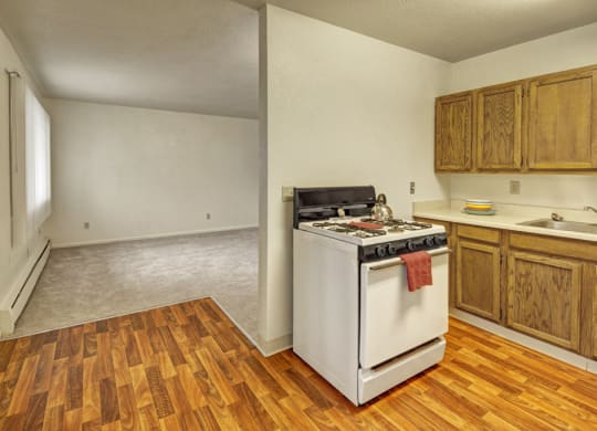 Small kitchen the Village at Calais Apartments with wooden cabinets, white stove, and wooden flooring. Adjacent is a carpeted living area; both areas are empty, creating a neutral, simple tone.