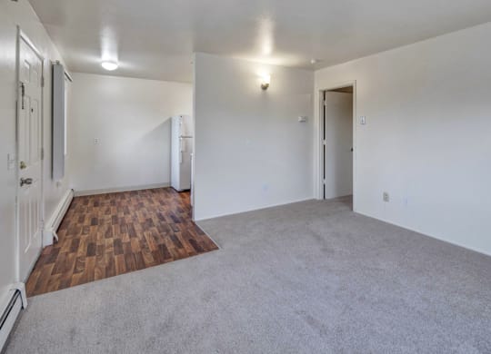 Empty living room here at the Village at Calais Apartments with light gray carpet and white walls, transitioning into a small kitchen area with wood-patterned flooring. Minimalist and clean.