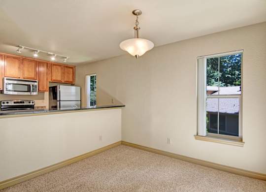 A neutral dining area here at Asbury Park featuring soft carpet flooring, a centered pendant light, and an open pass-through to the kitchen with a dark countertop, warm wood cabinetry, and stainless-steel appliances, while tall windows with vertical blinds bring in natural light and offer views of surrounding greenery.