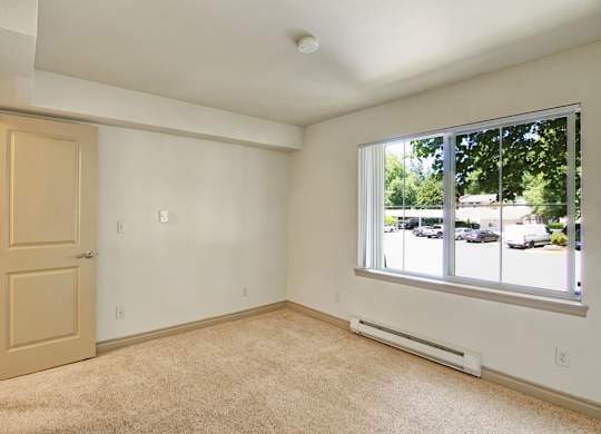 A neutral bedroom here at Asbury Park featuring plush carpet flooring, soft-toned walls, and a wide window with vertical blinds that brings in natural light and offers views of the community parking area, complemented by a simple door entry, clean trim details, and a calm layout ideal for rest and personalization.