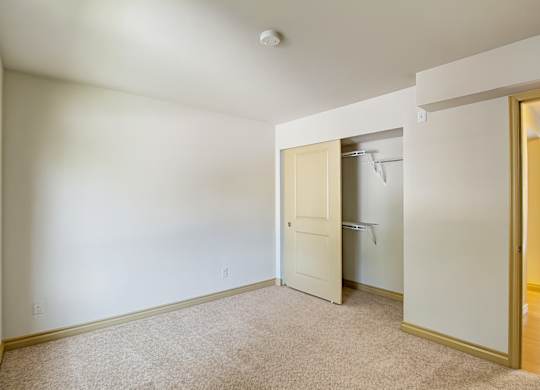 A comfortable bedroom here at Asbury Park featuring plush carpet flooring, soft neutral walls, and a large window that allows natural light to brighten the space, along with an open closet with shelving, a partially open interior door leading to the hallway, and ample wall space suitable for a bed, dresser, or additional storage.