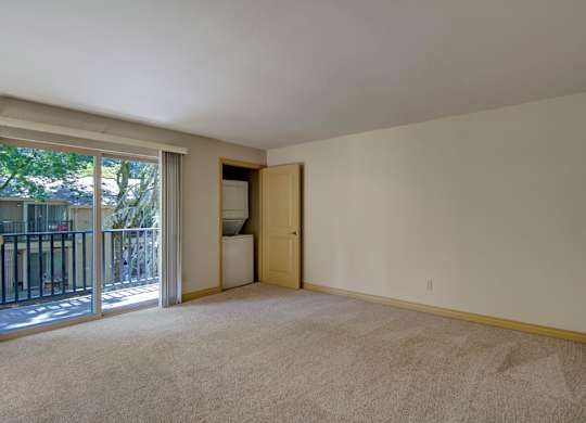 A spacious living room here at Asbury Park featuring plush carpet flooring, soft neutral walls, and a large sliding glass door that opens to a private balcony overlooking mature trees, along with an in-home laundry closet tucked behind a partially open door and an open layout offering ample wall space for seating and entertainment furniture.