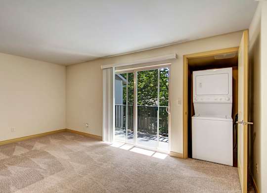 A spacious living room here at Asbury Park featuring soft neutral walls, plush carpet flooring, and a sliding glass door that opens to a private balcony with railing and leafy outdoor views, while a dedicated in-home laundry closet with stacked appliances is conveniently tucked behind a nearby door, offering a functional open layout with ample natural light.