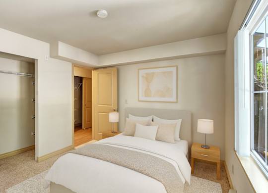 A cozy, well-lit bedroom here at Asbury Park featuring a neatly made bed with white bedding and soft neutral accent pillows, a light upholstered headboard, and matching bedside tables with modern lamps, complemented by plush carpet, an open closet with shelving, and a large window that brings in natural light.