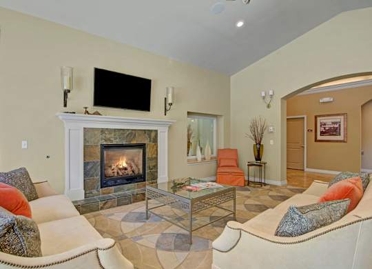 A welcoming resident lounge here at Asbury Park featuring a cozy seating area arranged around a tiled fireplace with a decorative mantel, a wall-mounted television, plush sofas with patterned accent pillows, a glass coffee table on a circular area rug, warm neutral walls, soft lighting, and an adjacent hallway that adds to the open community feel.