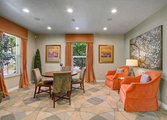 A comfortable resident lounge here at Asbury Park featuring a round dining table with upholstered chairs, two plush accent armchairs with a side table and lamp, patterned carpet flooring, framed wall art, soft neutral walls, ceiling lighting, and large windows with drapery that bring in natural light and create a relaxed gathering space.