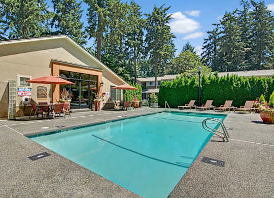 A sunlit outdoor swimming pool here at Asbury Park featuring a clear rectangular pool with metal handrails, a wide textured pool deck, lounge chairs along the edge, shaded patio tables with umbrellas, and a clubhouse building with large windows, all surrounded by tall evergreens and manicured hedges creating a relaxed community setting.