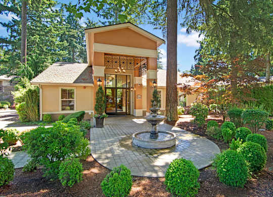 A welcoming leasing office entrance here at Asbury Park showcasing a charming building with a covered entry, glass front doors, and warm exterior accents, centered around a decorative fountain on a paved walkway, surrounded by manicured shrubs, mature trees, and landscaped greenery that create an inviting, park-like community setting.