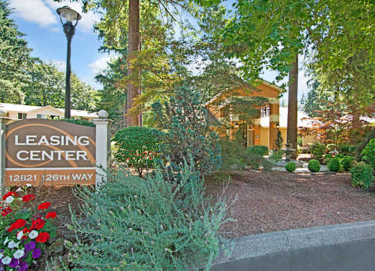 A landscaped leasing center entrance here at Asbury Park featuring a prominent leasing center sign along a curved driveway, surrounded by colorful flowers, manicured shrubs, and mature trees, with the leasing office building visible in the background, warm exterior lighting, and a tranquil fountain creating a welcoming, garden-style community atmosphere.