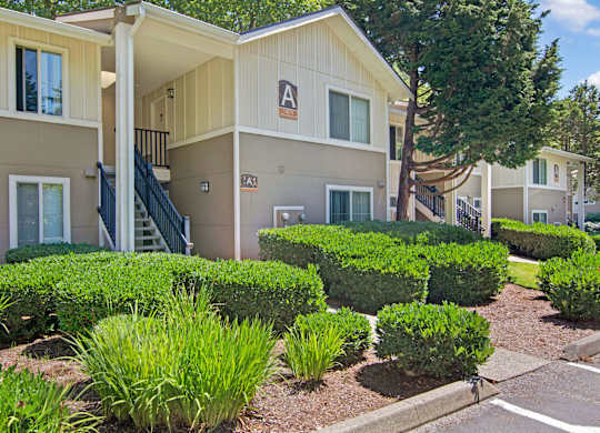 A well-maintained apartment building exterior here at Asbury Park featuring light-toned siding with contrasting trim, covered staircases with railings, and large windows, surrounded by neatly trimmed hedges, landscaped greenery, and mature trees, creating a clean, residential setting with clearly marked building signage and walkable paths.
