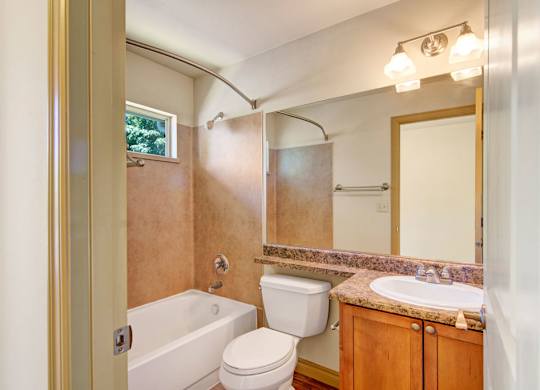 A clean, functional bathroom here at Asbury Park featuring a tub and shower combination with a curved curtain rod, a small window for natural light, and neutral wall surrounds, complemented by a granite-style vanity countertop with an undermount sink, wood cabinetry, a large wall mirror, and overhead lighting for everyday convenience.