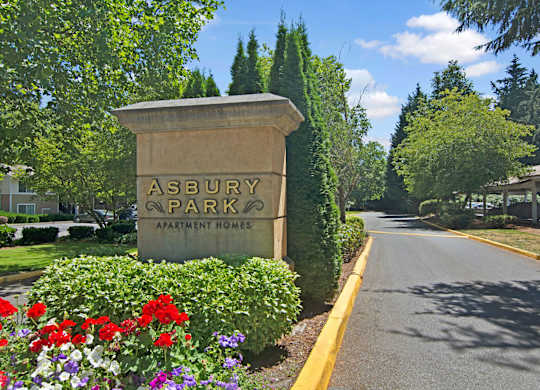 A landscaped community entrance sign here at Asbury Park featuring a stone monument displaying the property name, surrounded by colorful flowers, trimmed hedges, and mature trees, with a paved driveway extending into the community and covered parking visible in the background under a clear blue sky.