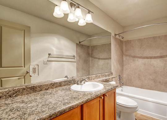 A well-appointed bathroom here at Asbury Park showcasing a spacious granite-style vanity countertop with an undermount sink, warm wood cabinetry, and a wide wall mirror beneath bright overhead lighting, paired with a tub and shower combination featuring neutral wall surrounds, a curved shower rod, and coordinated fixtures for a clean, practical layout.