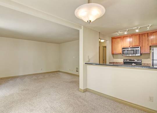 A spacious open living area here at Asbury Park featuring plush carpeted flooring, neutral walls, and a wide layout that flows into an open kitchen with warm wood cabinetry, a breakfast bar, a built-in microwave, and a smooth countertop, while ceiling lighting and track lights create a bright, inviting atmosphere with flexible space for living and dining.