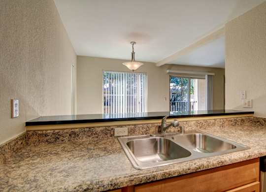 A welcoming kitchen pass-through here at Asbury Park showcasing smooth, speckled countertops with a double-basin sink and chrome faucet, opening to a dining and living area with vertical blinds, a sliding glass door, and a pendant light that adds warmth while allowing natural light to flow through the connected space.