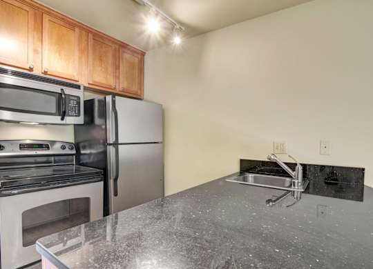 A modern kitchen here at Asbury Park featuring warm wood cabinetry, a smooth dark countertop with an integrated sink and sleek faucet, and stainless-steel appliances including a refrigerator, microwave, and range, with track lighting overhead that brightens the workspace and highlights the clean, functional layout.