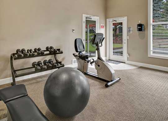 A functional fitness room here at Colonial Square featuring a rack of free weights, a stationary bike, and a stability ball set on neutral carpeting, with large windows and glass doors bringing in natural light, light-colored walls, and open floor space that creates a simple and practical workout environment.