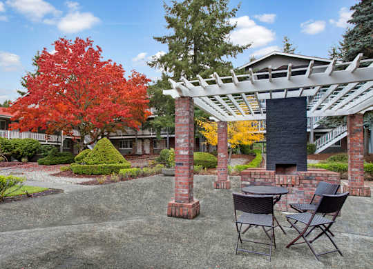 A landscaped outdoor courtyard here at Colonial Square featuring a brick pergola with a built-in grill, a small round table with chairs, and a paved seating area surrounded by manicured shrubs, gravel pathways, and vibrant red and yellow autumn trees, with two-story apartment buildings and exterior walkways in the background.