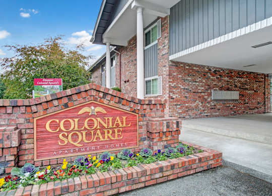 A welcoming monument sign here at Colonial Square displaying the community name set into a red brick base with colorful landscaping, positioned beside a covered building entry featuring brick exterior walls, white columns, gray siding accents, and a paved walkway leading toward the apartment homes.