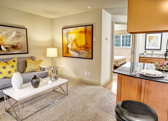 A cozy open living area here at Colonial Square showcasing a comfortable gray sofa with yellow accent pillows, a modern coffee table with decorative accents, and warm carpeting, flowing into a compact kitchen with wood cabinetry, dark countertops, a breakfast bar with seating, and a view toward a bedroom and vanity area that creates a connected, inviting layout.