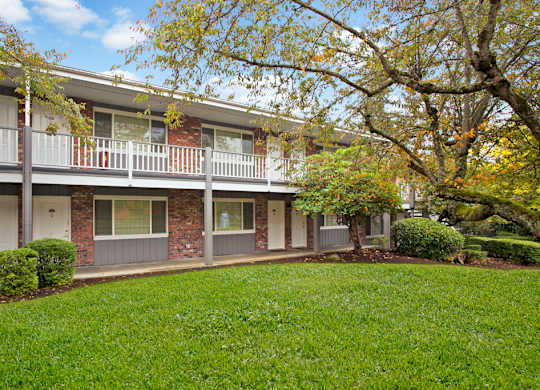 A tree-lined exterior courtyard here at Colonial Square featuring two-story brick apartment buildings with white railings and exterior walkways, ground-level entry doors and windows, and a well-maintained green lawn bordered by shrubs and mature trees with early autumn foliage under a bright blue sky.