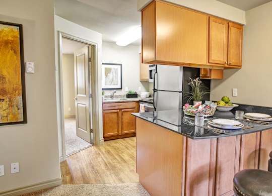A compact kitchen here at Colonial Square featuring warm wood cabinetry, dark countertops with bar seating, and a small breakfast bar set with plates and glassware, complemented by wood-style flooring, a stainless-look refrigerator, a nearby sink area with additional cabinets, and soft overhead lighting that creates a functional and welcoming cooking space.