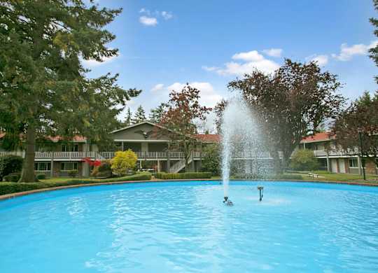 A serene fountain courtyard here at Colonial Square showcasing a large circular pool with a tall central water feature, surrounded by mature evergreen and deciduous trees, manicured hedges, and open green lawn areas, with two-story apartment buildings and exterior walkways framing the peaceful, park-like community setting.