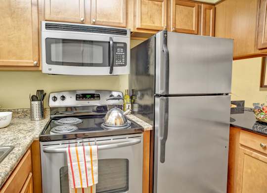 A compact kitchen here at Colonial Square featuring warm wood cabinetry, speckled countertops, a stainless refrigerator, a built-in microwave above a smooth cooktop range, and a sink set into the counter, with simple decor and neatly arranged cookware creating a functional, efficient space for everyday cooking and meal prep.