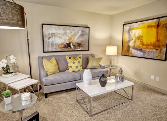 A cozy living room here at Colonial Square featuring a soft gray sofa with yellow and patterned accent pillows, a sleek white coffee table with decorative vases, and a side table with a modern lamp, complemented by abstract wall art, neutral carpeting, and warm ambient lighting that creates an inviting and comfortable atmosphere.
